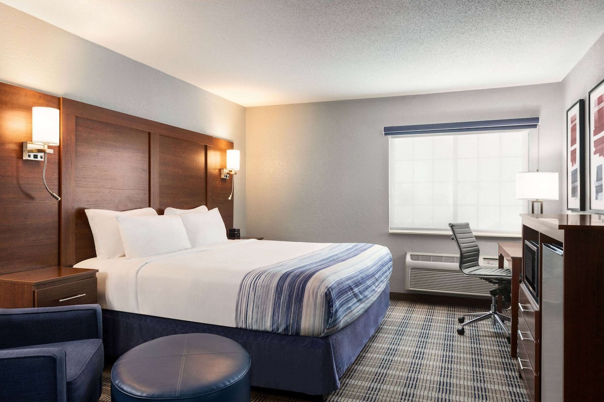 A hotel room with a large bed, a desk with a chair, a small table, and a window with blinds.
