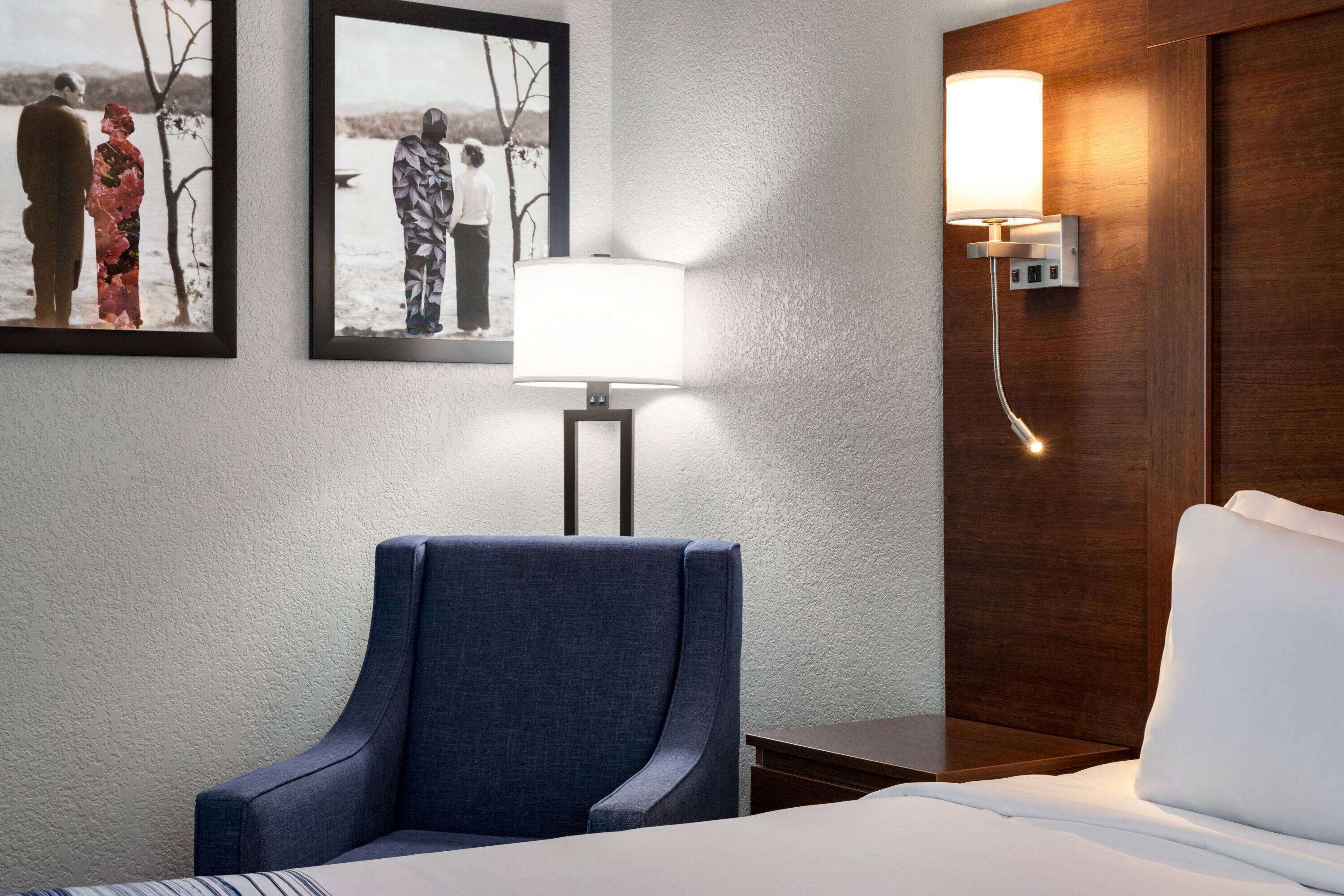 A hotel room with a bed, a blue armchair, a wall-mounted lamp, and two framed photographs on the wall.