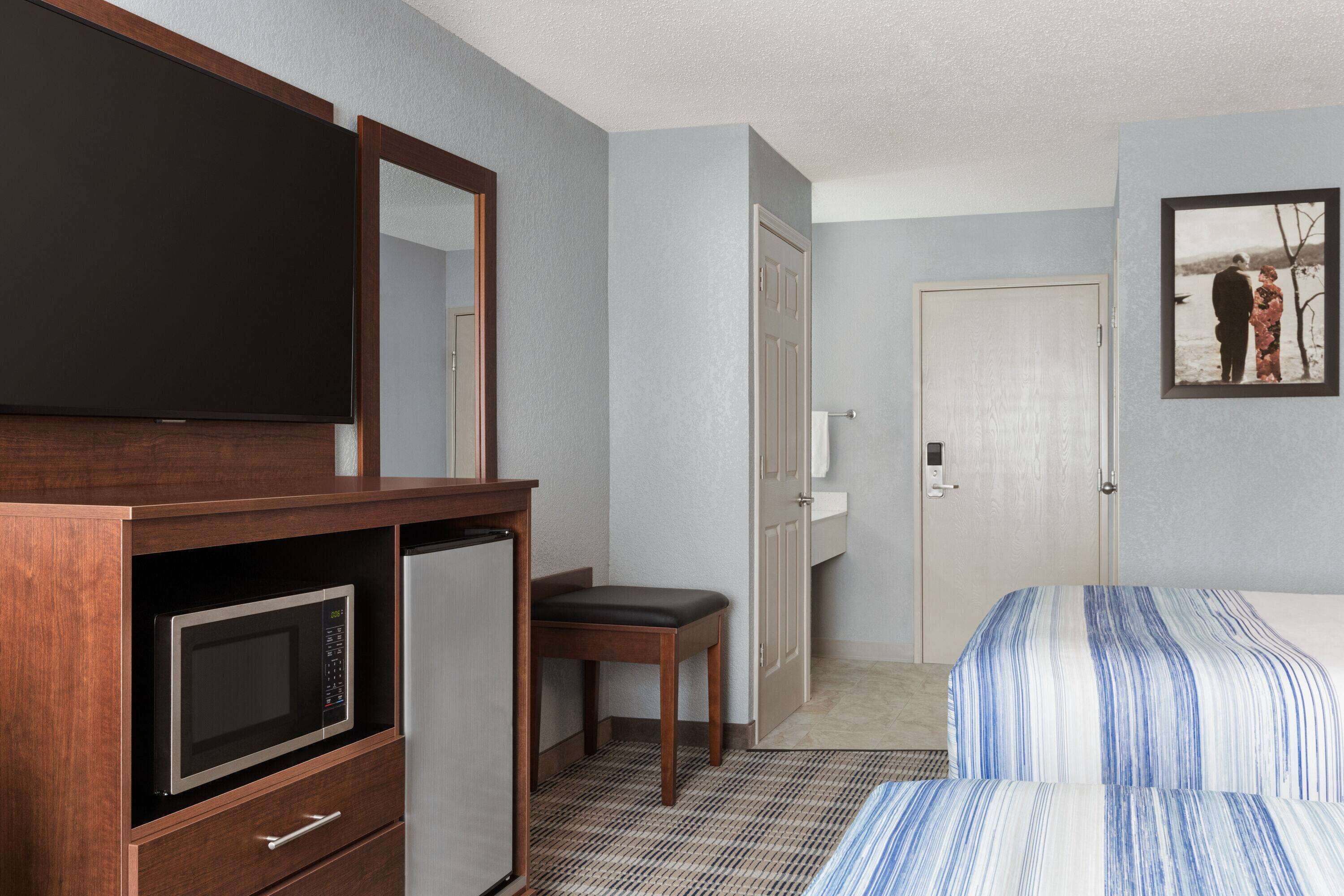 Habitación de hotel con una cama, un escritorio con microondas, una silla, un televisor y una puerta que da a un baño.