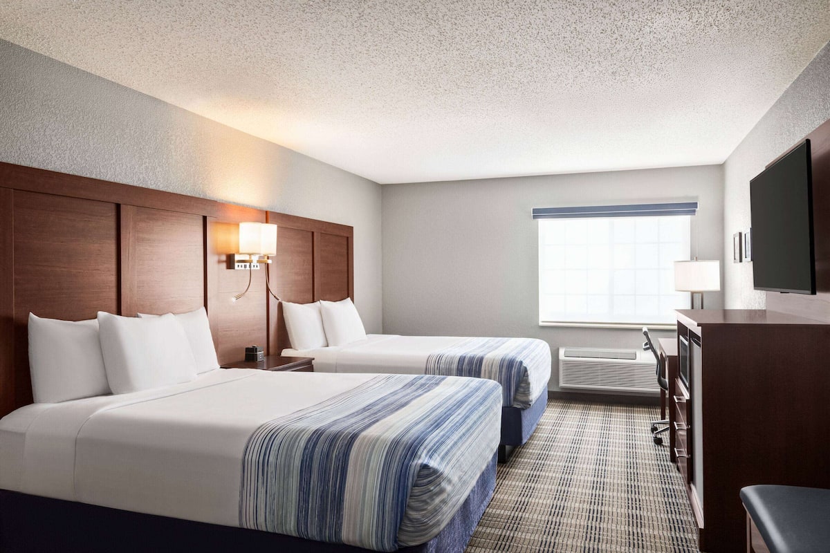 A hotel room with two beds, a wooden headboard, a flat-screen TV, a desk, and a window with blinds.