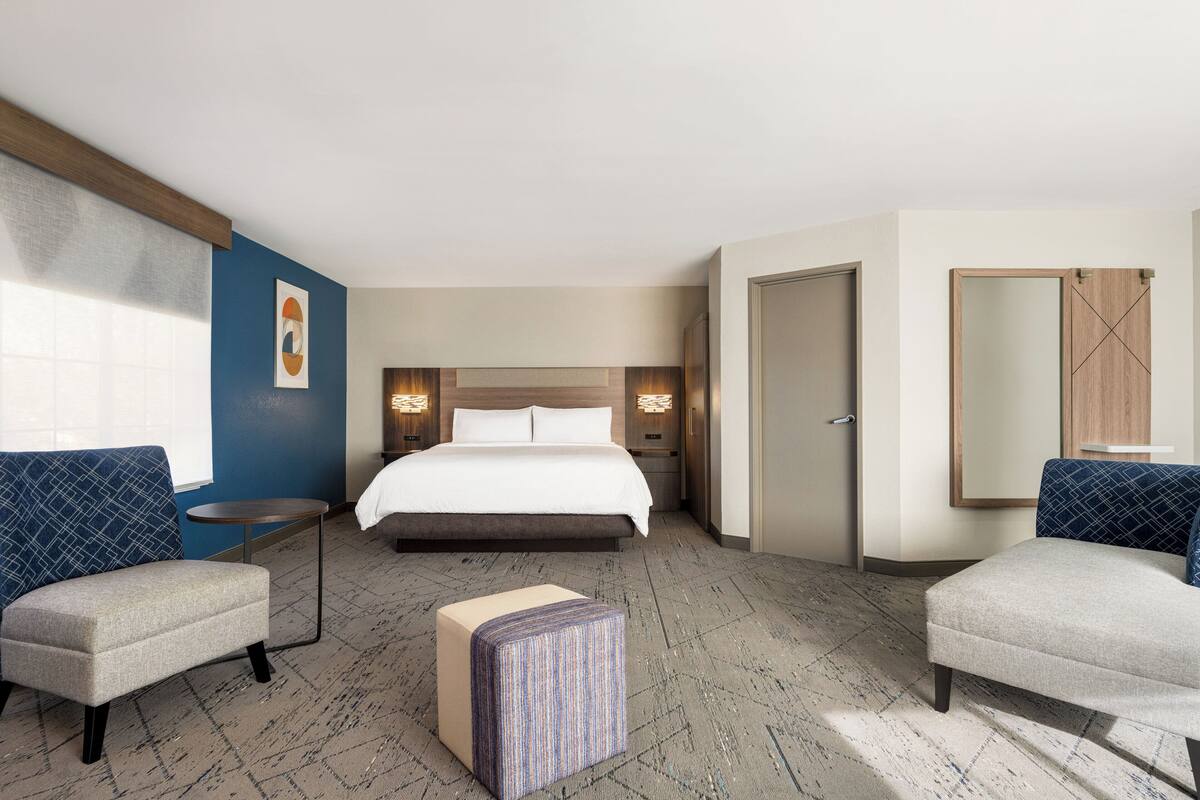 A hotel room with a bed, two armchairs, a small table, a mirror, and a door.