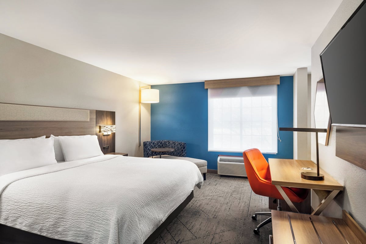 A hotel room with a large bed, a desk with a chair, a small table, and a window with blinds.