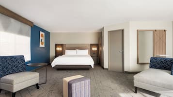 Habitación de hotel con una cama, dos sillones, una mesita, una mesita de noche, un espejo y un cuadro geométrico en la pared.
