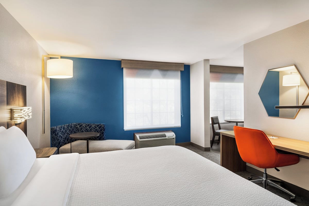 A hotel room with a bed, a desk, a chair, a small table, and a window with blinds.