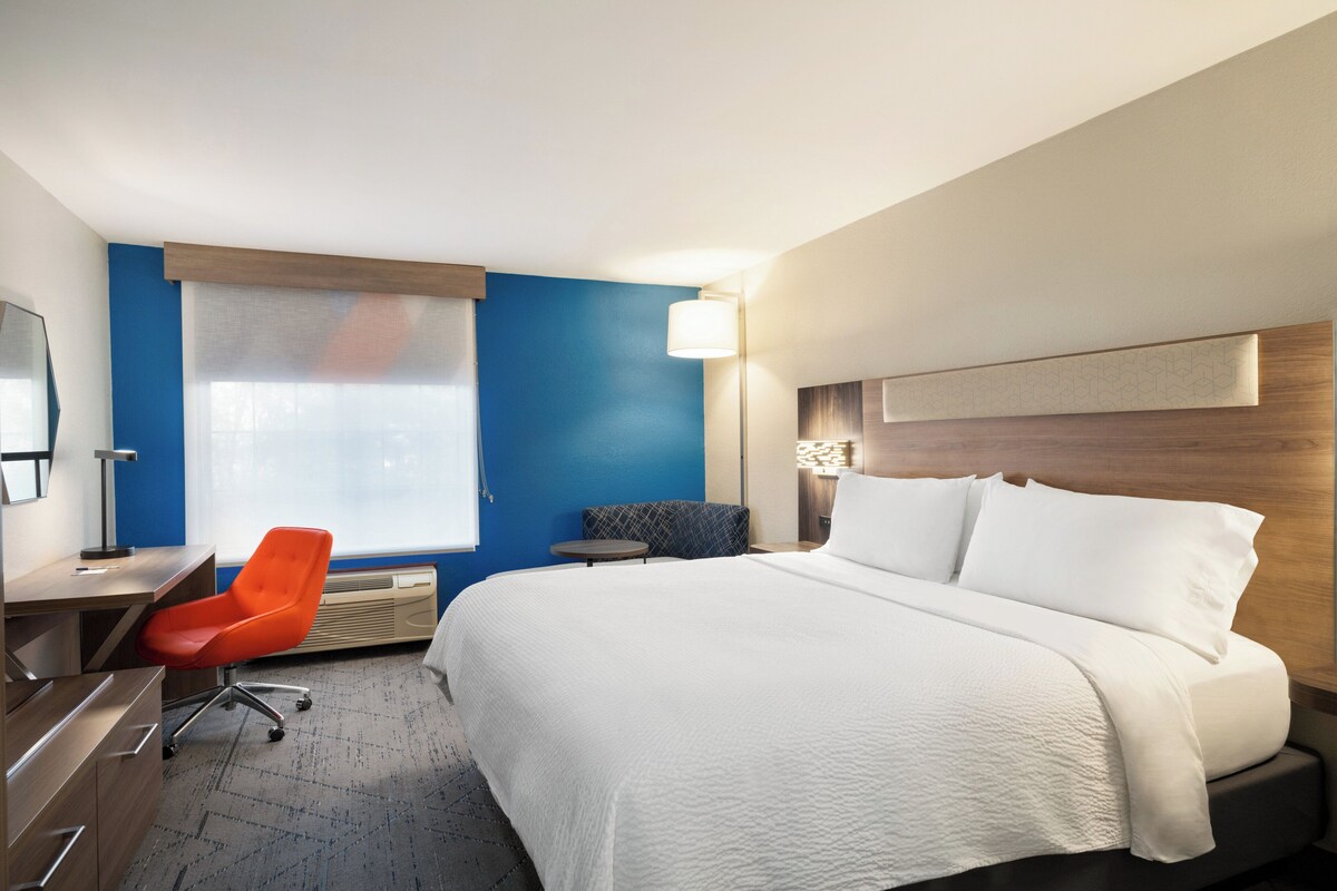 Ein Hotelzimmer mit einem großen Bett, einem Schreibtisch mit Stuhl, einem kleinen Tisch und einem Fenster mit Jalousien.