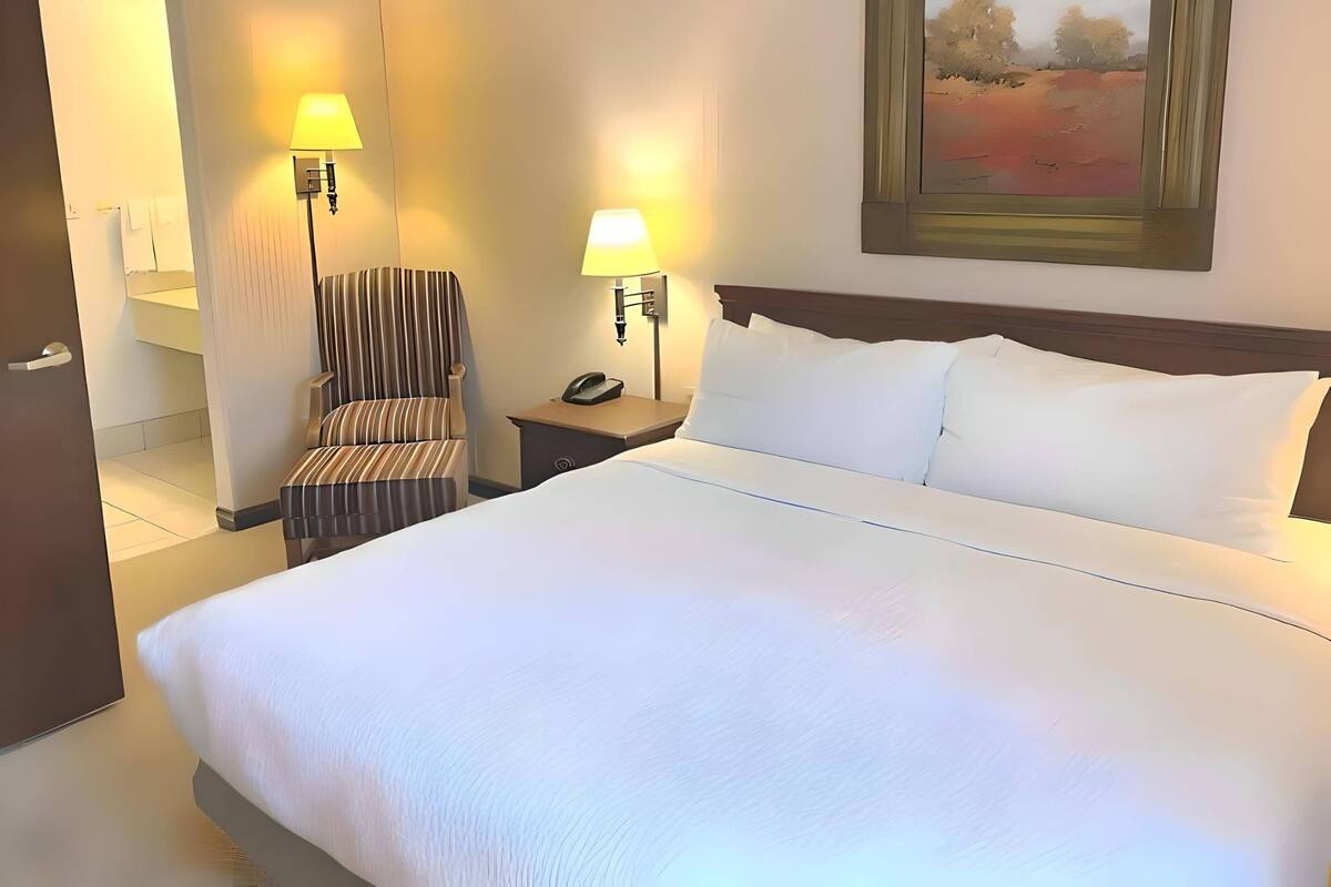Una habitación de hotel con una cama grande, dos lámparas de noche, una silla, una mesita de noche y un cuadro en la pared.