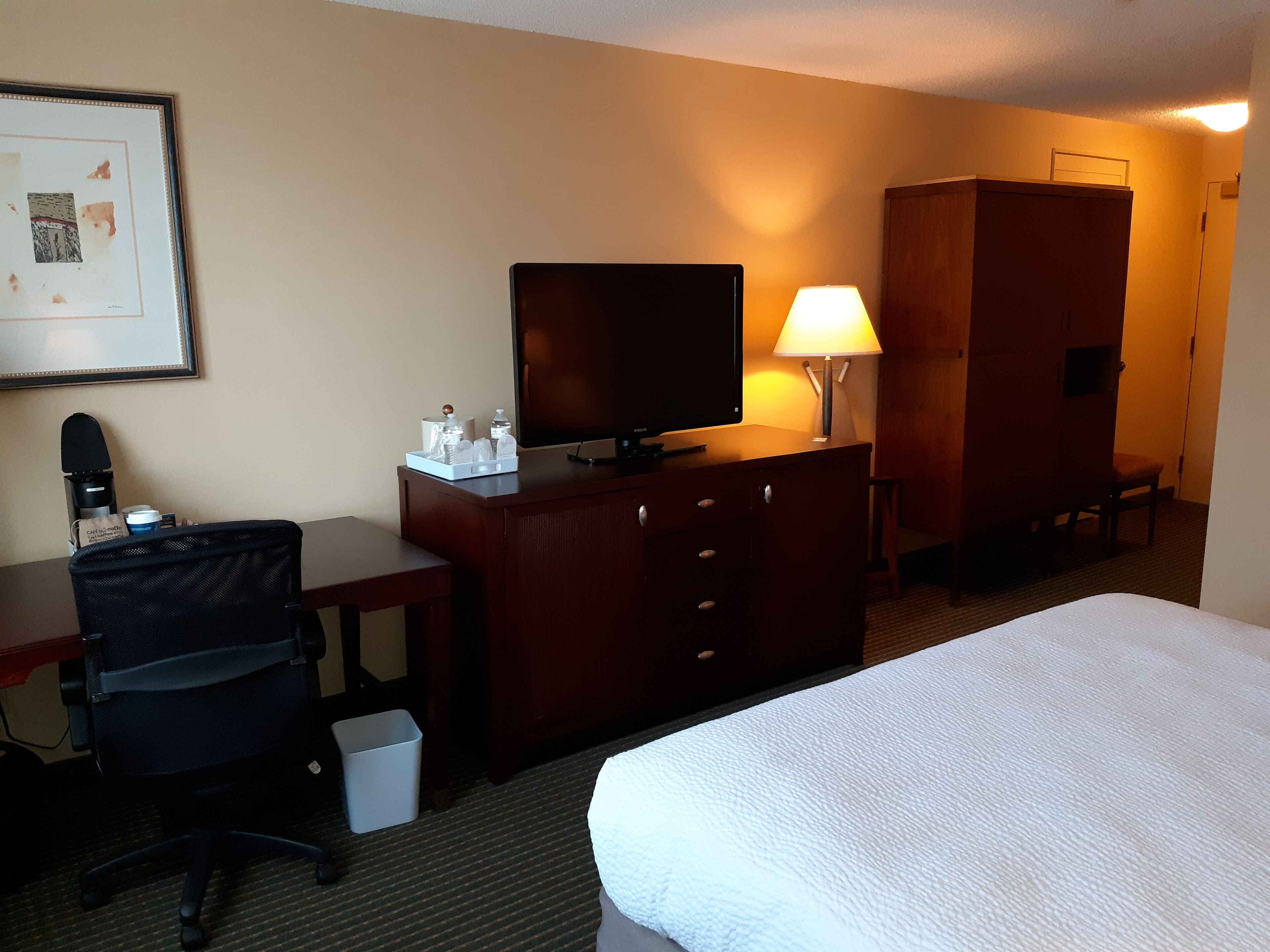 A hotel room with a bed, a desk with a chair, a television, a lamp, and a wardrobe.