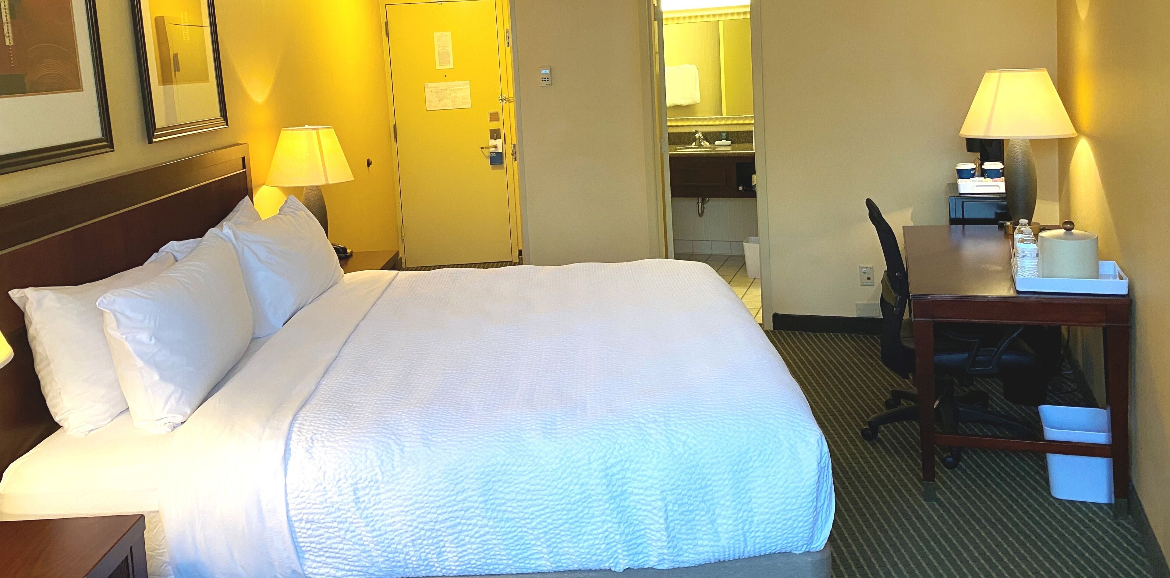 A hotel room with a large bed, two bedside lamps, a desk with a chair, and a small table with a lamp.
