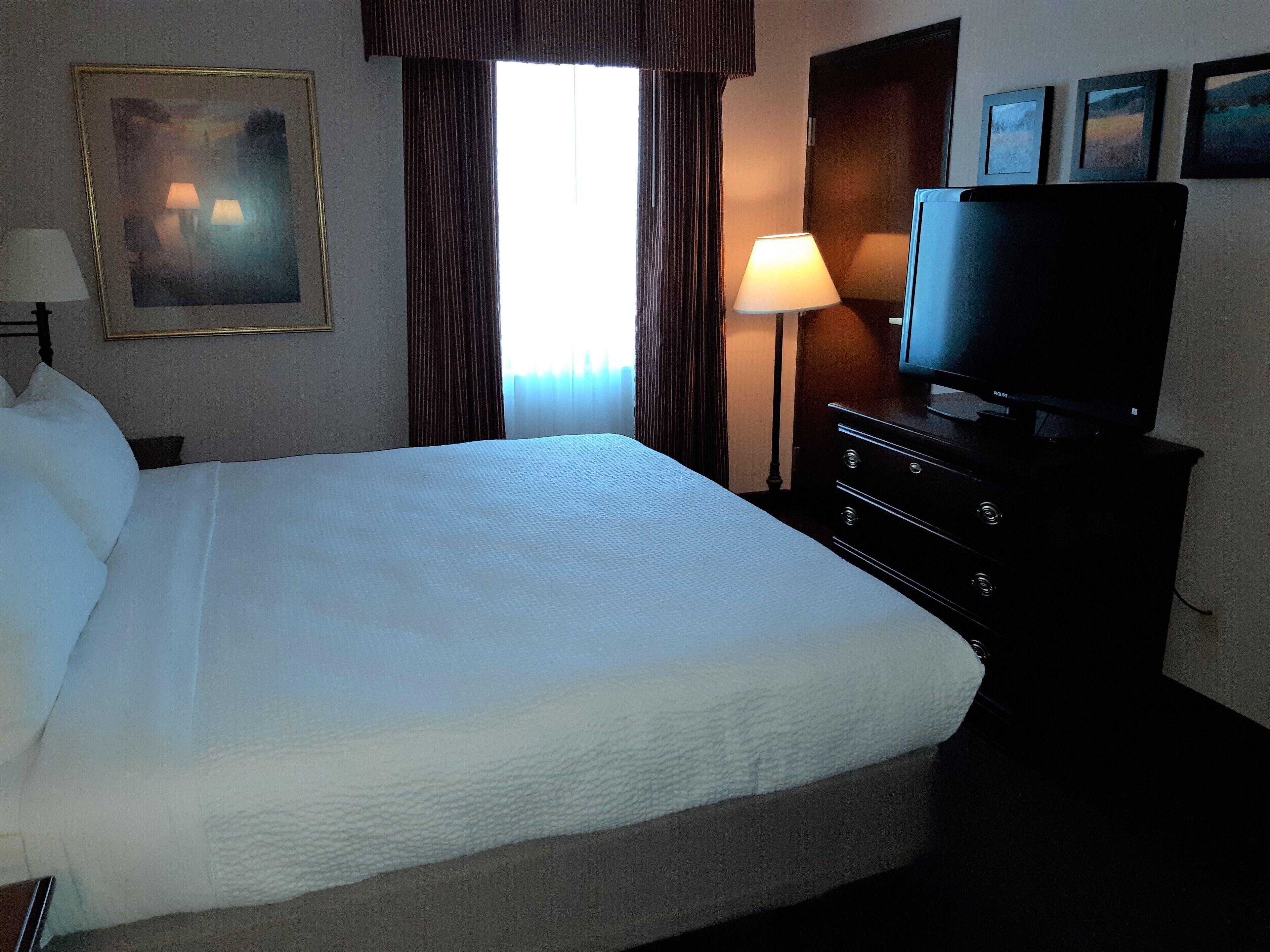 A hotel room with a large bed, a television on a dresser, a lamp, and a painting on the wall.