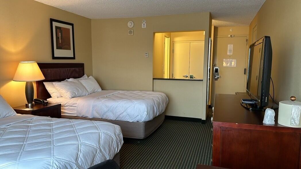 A hotel room with two beds, a desk with a television, a mirror, and a lamp.