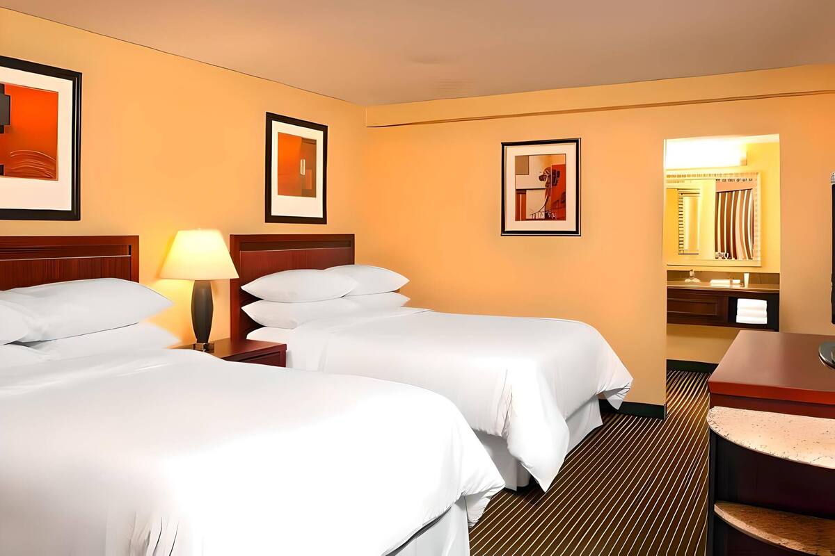 Habitación de hotel con dos camas, una mesita de noche con lámpara, espejo de baño y cuadros en las paredes.