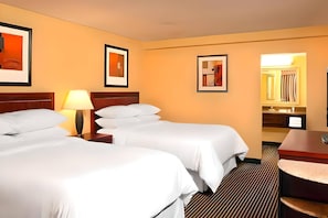 Quarto de hotel com duas camas, mesa de cabeceira com abajur, espelho no banheiro e quadros nas paredes.