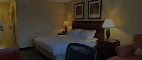 Una habitación de hotel con una cama, dos mesitas de noche, una silla, un escritorio y una puerta.