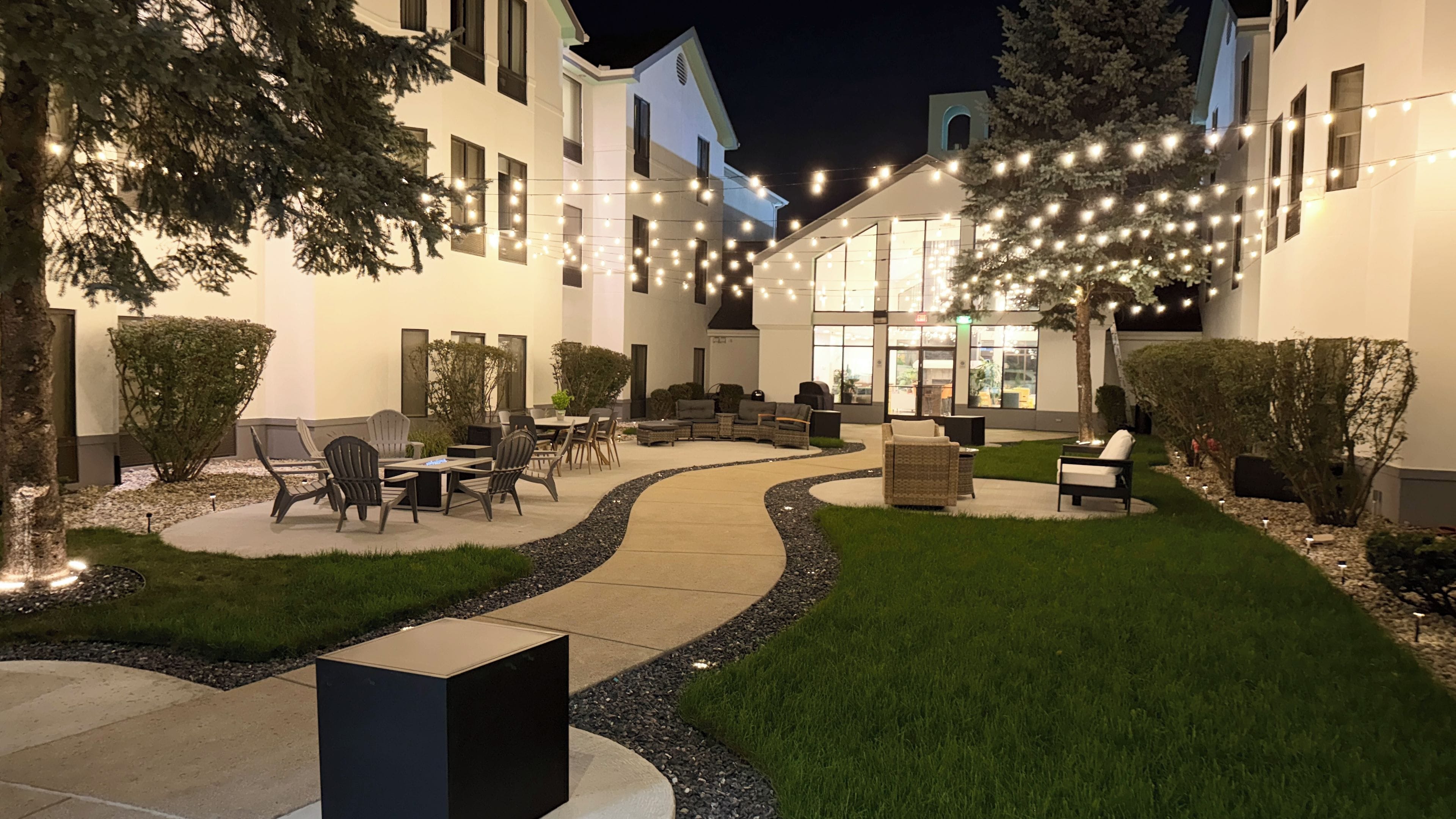 Ein gut beleuchteter Außenbereich mit Terrasse, Sitzgelegenheiten, einem Weg und einem Gebäude im Hintergrund.