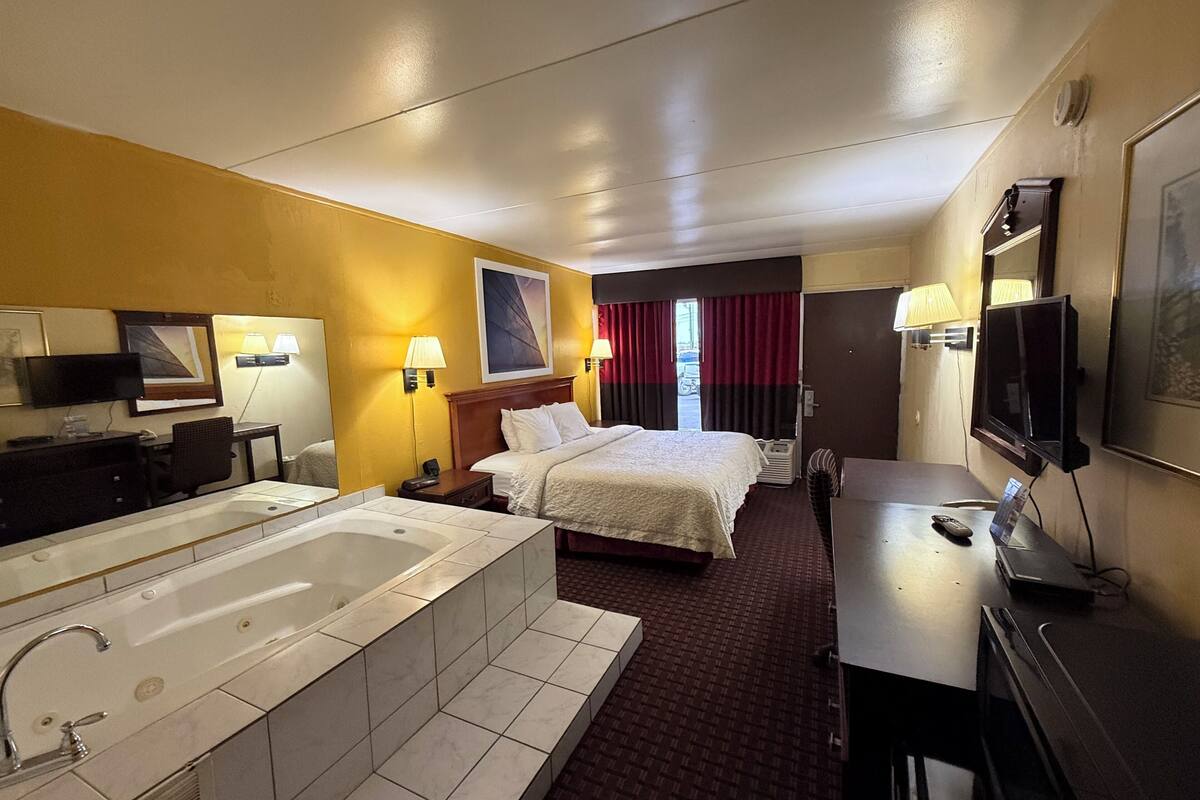 A hotel room with a large bed, a separate desk, a television, a bathroom with a bathtub, and a window with curtains.