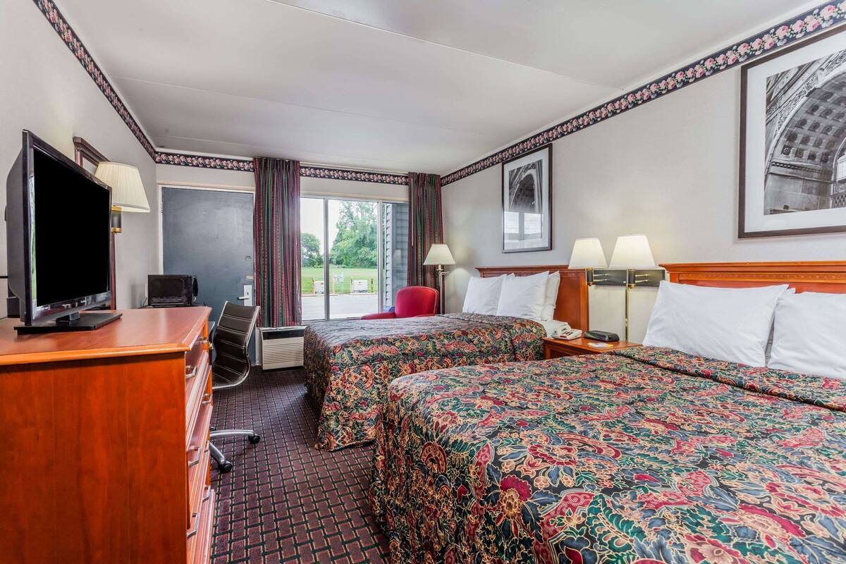 A hotel room with two beds, a desk with a TV, a chair, a red armchair, and a window with curtains.