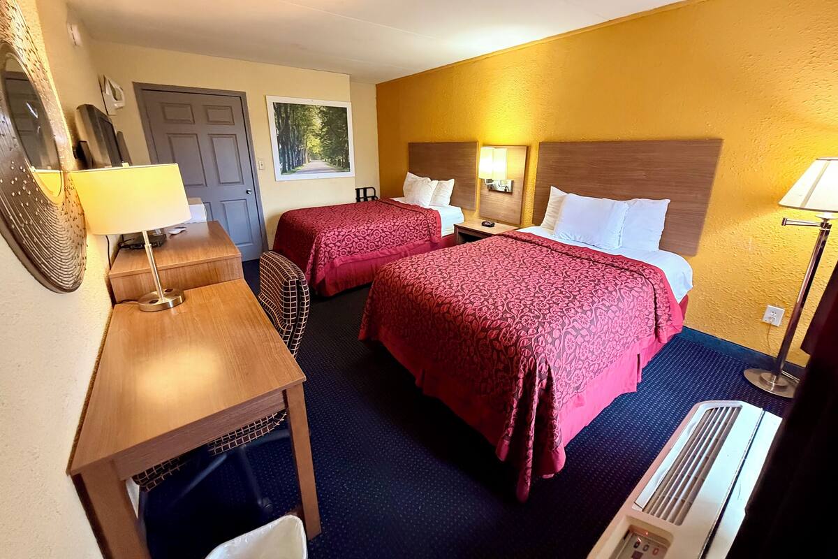 A hotel room with two beds, a desk, a chair, a heater, and a wall-mounted TV.