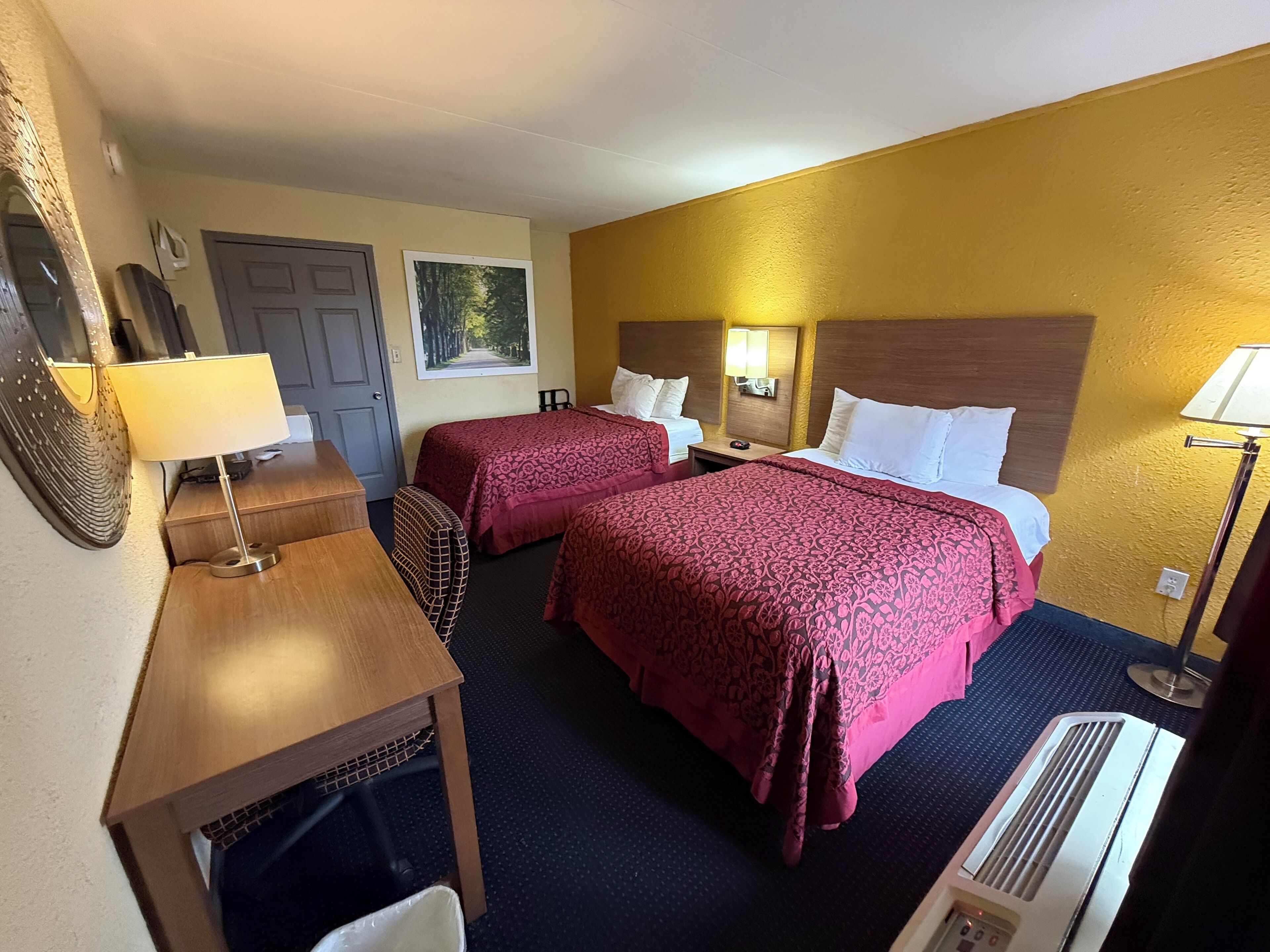 A hotel room with two beds, a desk, a chair, a lamp, and a heater.