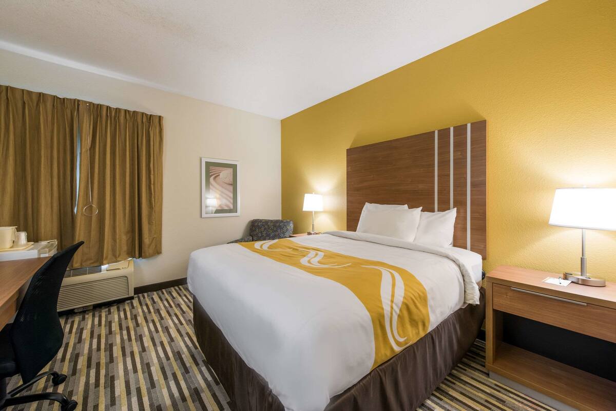 A hotel room with a large bed, a desk with a chair, a bedside table with a lamp, and a wall-mounted air conditioner.