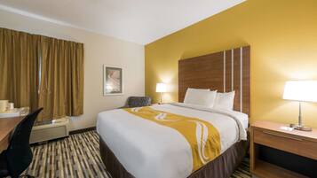 A hotel room with a large bed, a desk with a chair, a bedside table with a lamp, and a wall-mounted air conditioner.