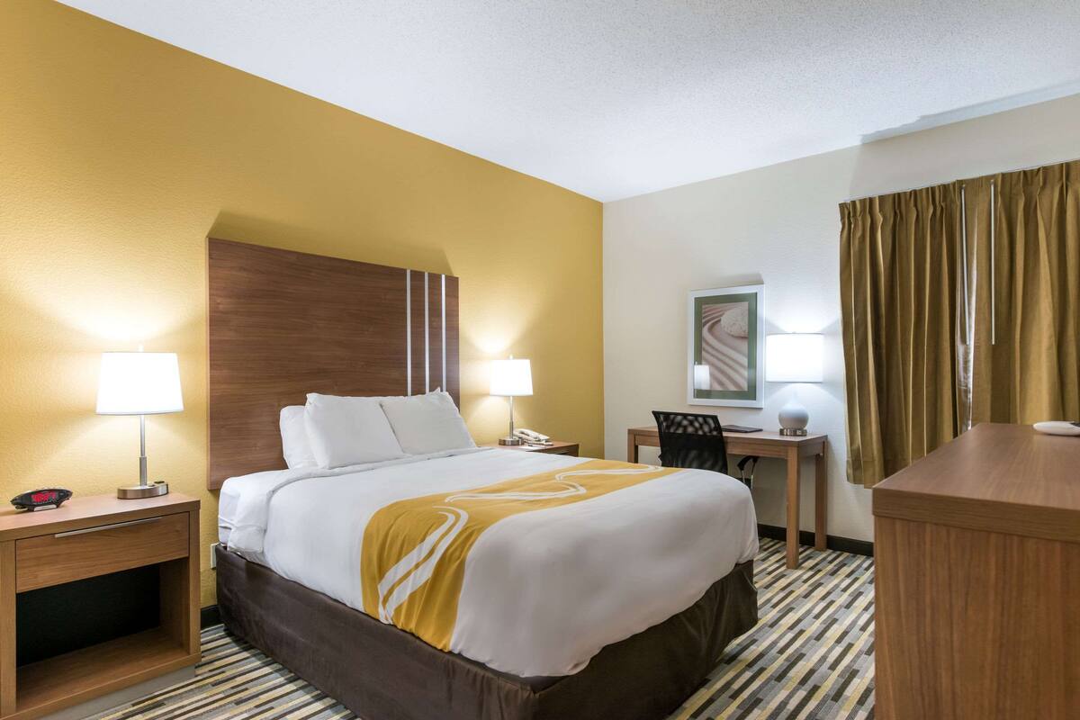 A hotel room with a bed, bedside lamps, a desk, a chair, and a mirror.