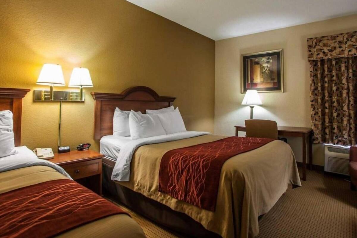 A hotel room with two beds, a desk, a chair, a lamp, a painting, and a window with curtains.