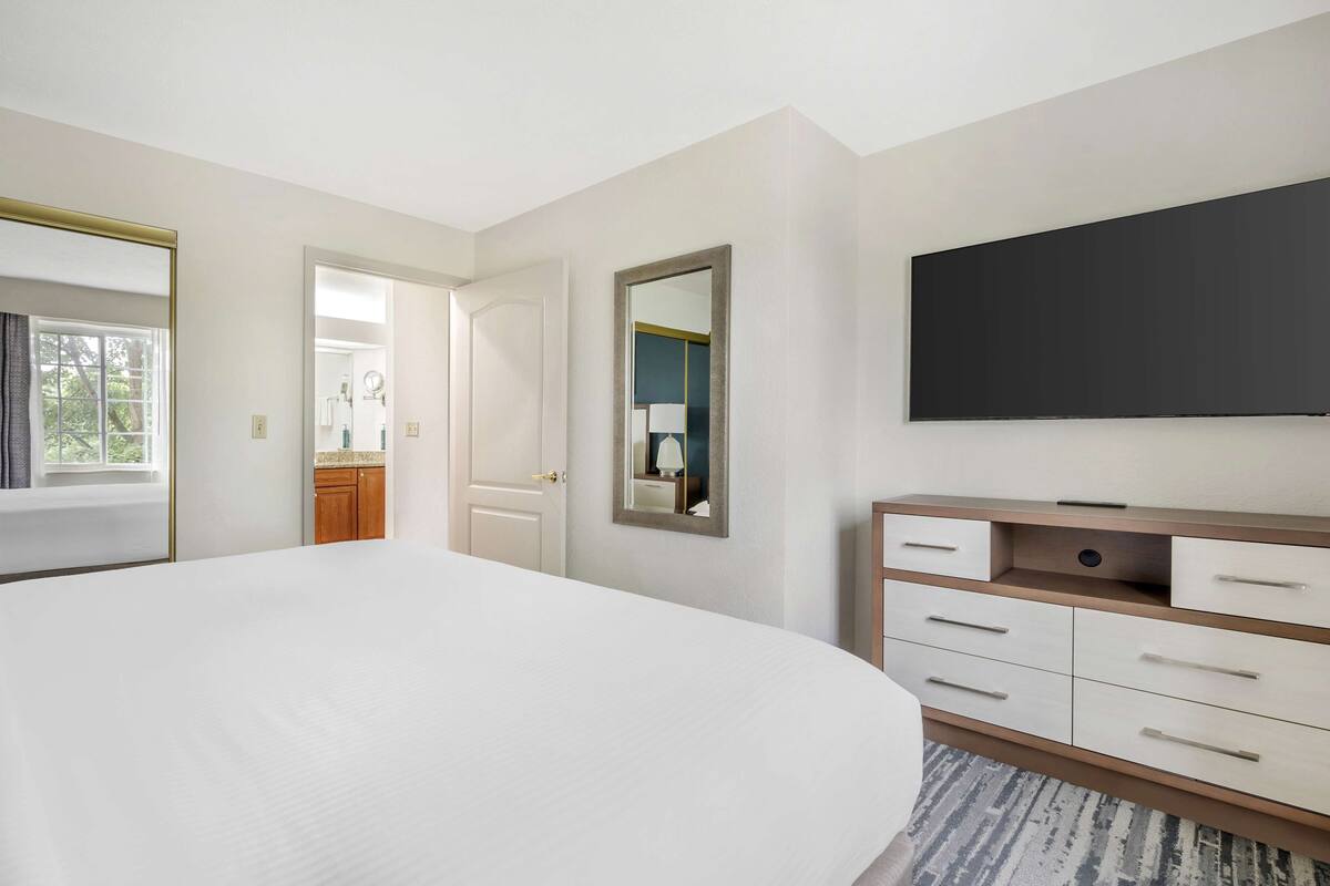 A bedroom with a large bed, a dresser, a mirror, and a flat-screen TV mounted on the wall.