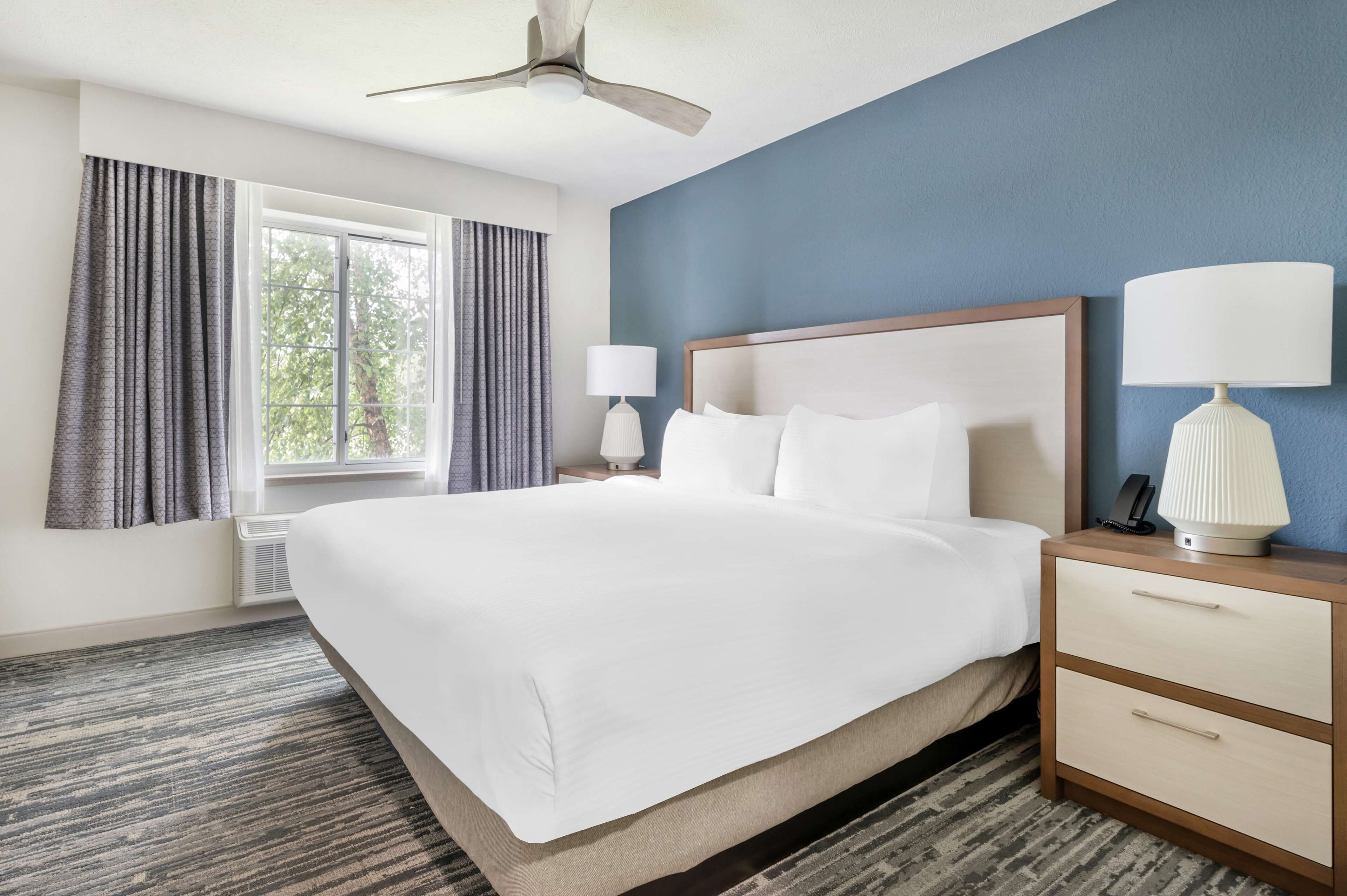 Une chambre d’hôtel avec un grand lit, une table de chevet avec une lampe, un ventilateur de plafond et une fenêtre avec des rideaux.