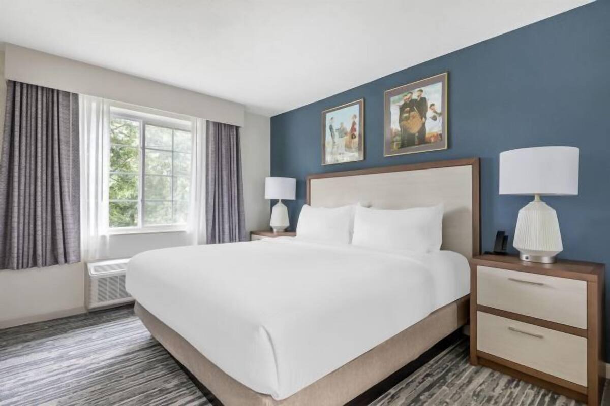 A hotel room with a large bed, two bedside tables with lamps, a window with curtains, and two framed pictures on the wall.