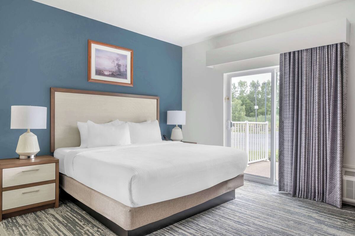 A hotel room with a large bed, two bedside tables with lamps, a framed picture on the wall, and a view of the outdoors through a sliding glass door.