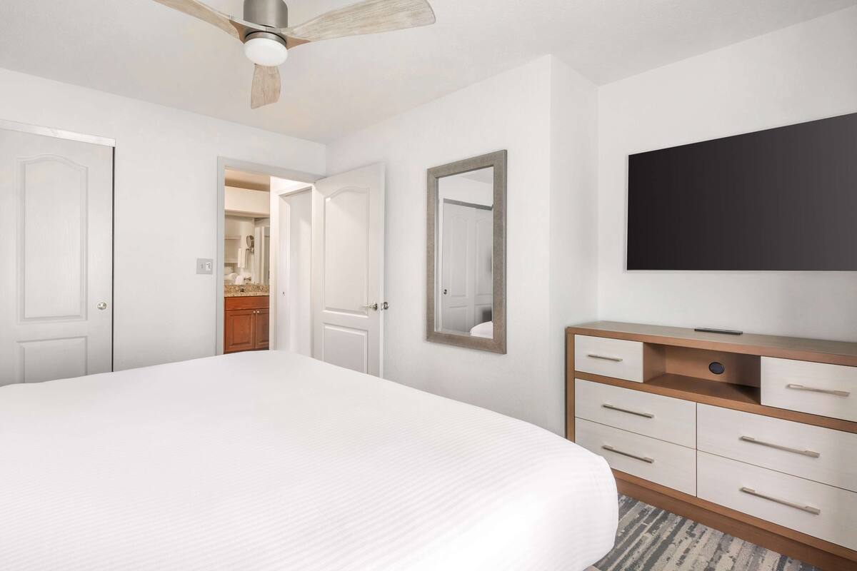 A hotel room with a large bed, a wooden dresser, a flat-screen TV, and a ceiling fan.