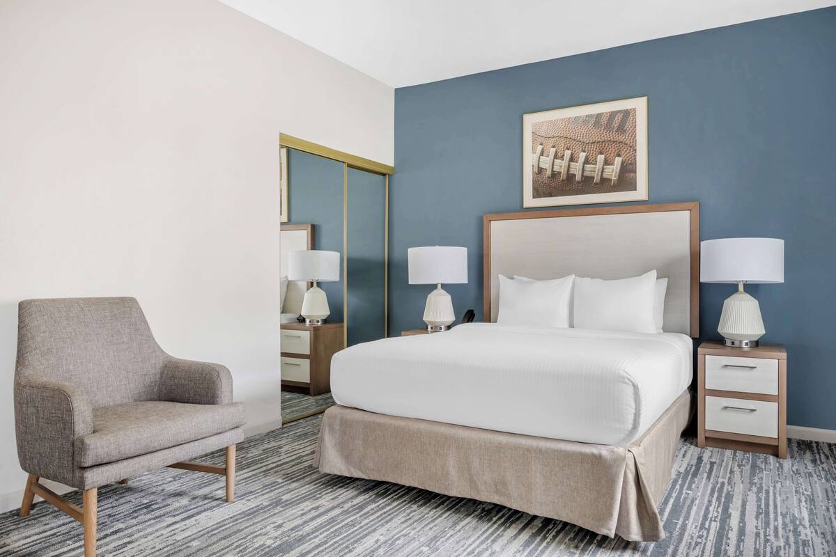 A hotel room with a bed, a chair, two bedside tables with lamps, a mirror, and a framed picture on the wall.