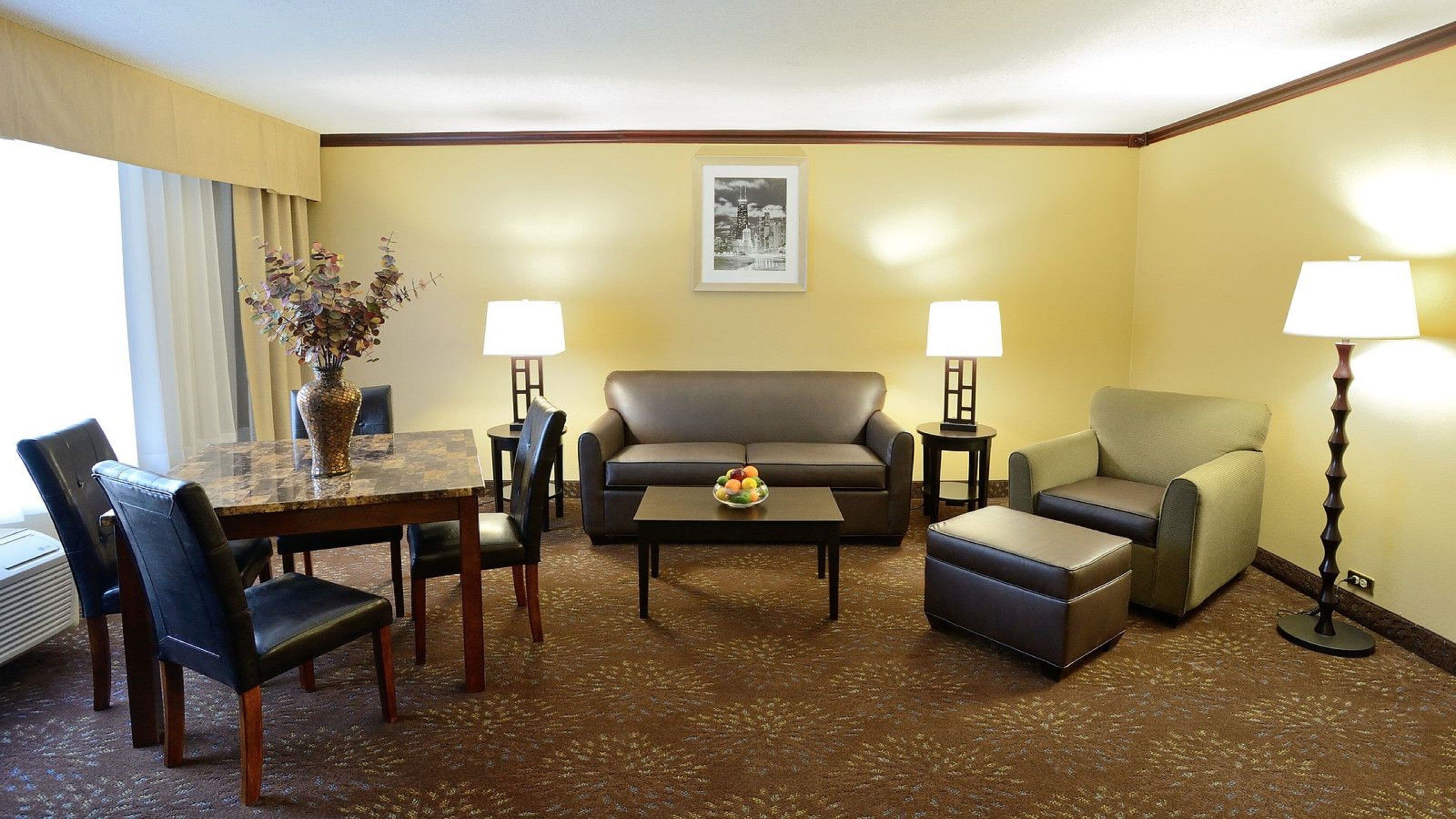 A hotel room with a dining table, two chairs, a sofa, a loveseat, a coffee table, and two lamps.