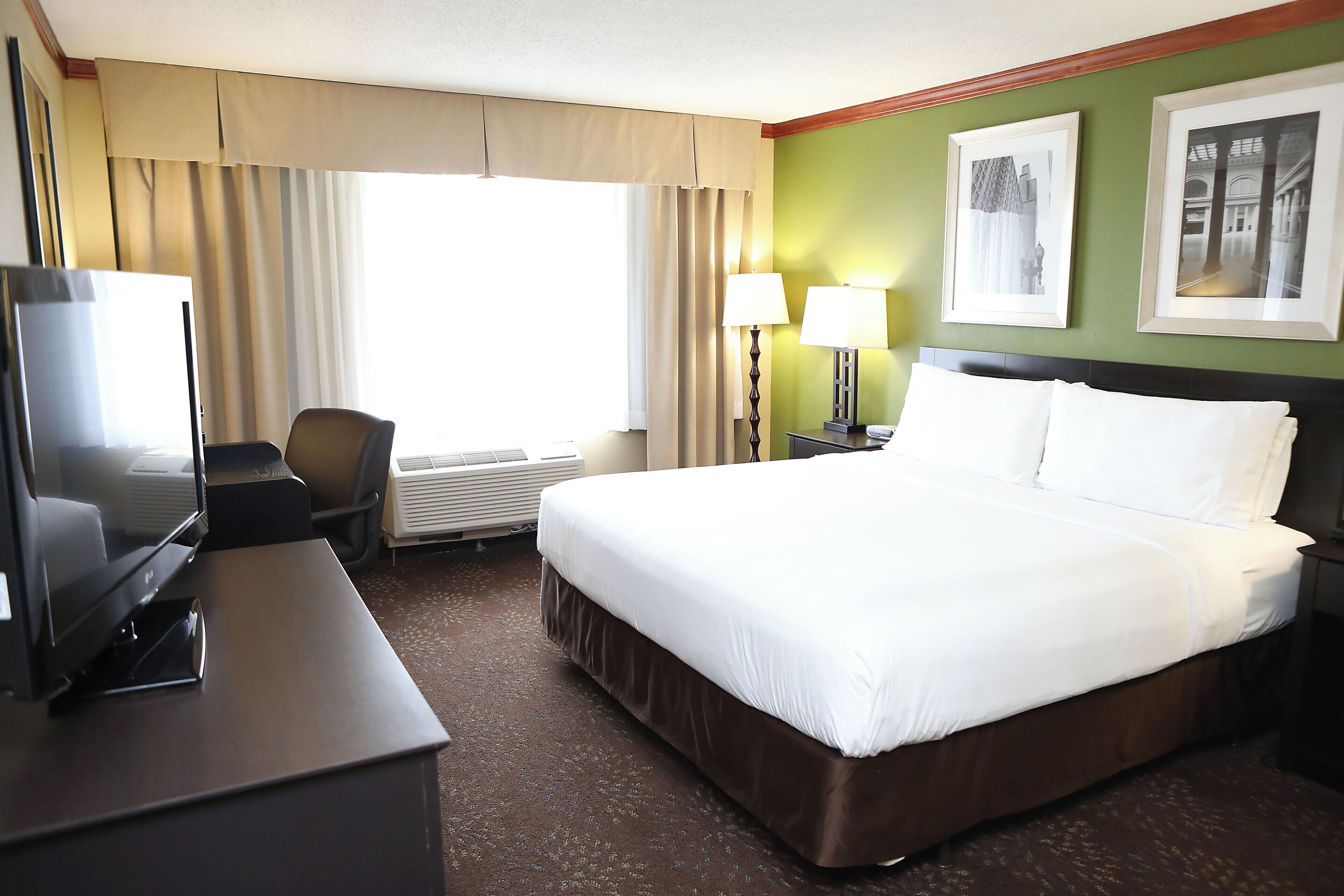 Habitación de hotel con cama, escritorio con televisor, silla, lámpara y ventana con cortinas.