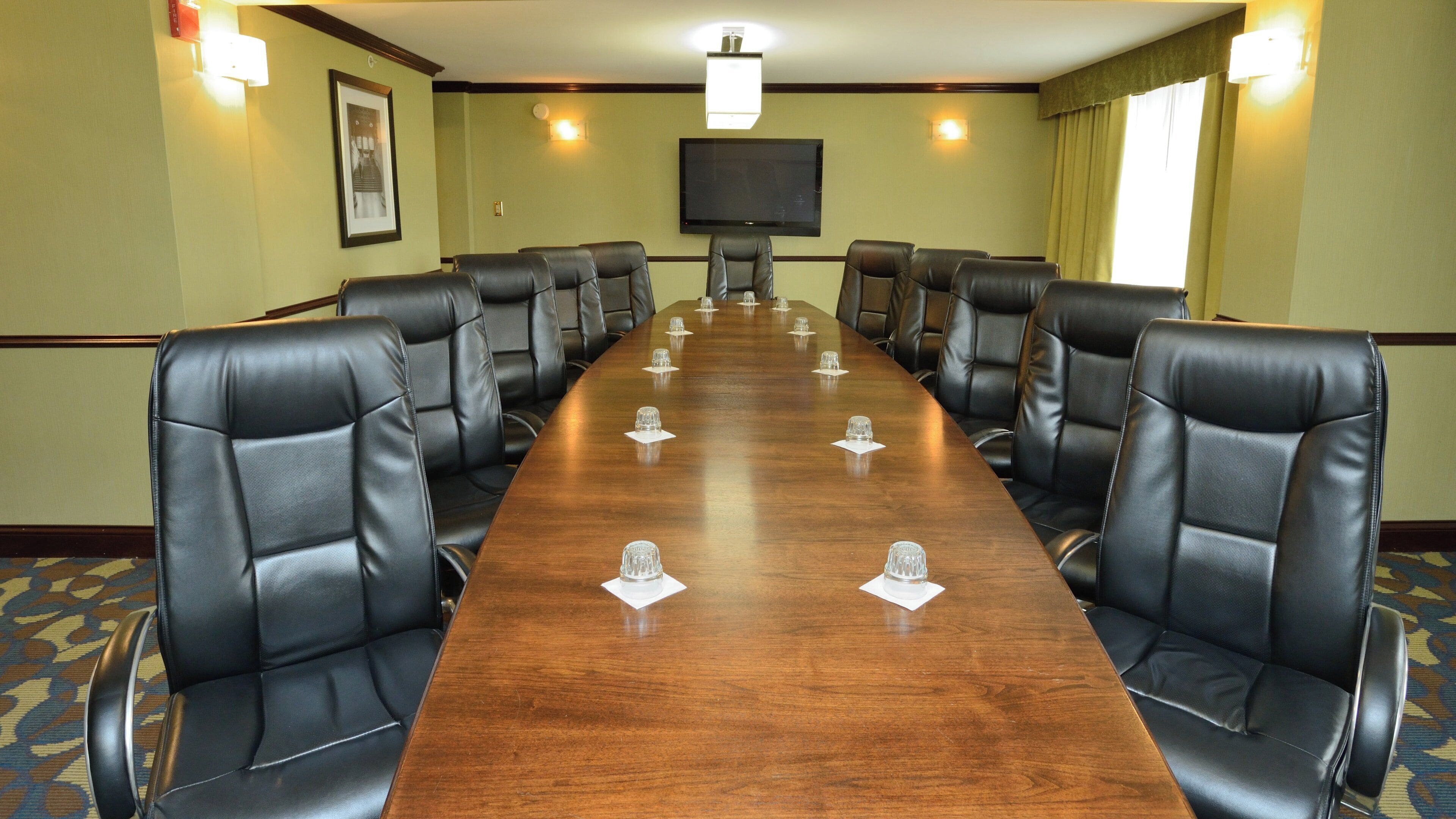 A conference room with a long wooden table, black leather chairs, a flat-screen TV, and a framed picture on the wall.