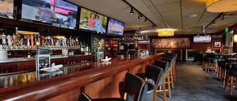 Bar com várias telas exibindo eventos esportivos, diversas torneiras de cerveja e área de estar com banquetas.