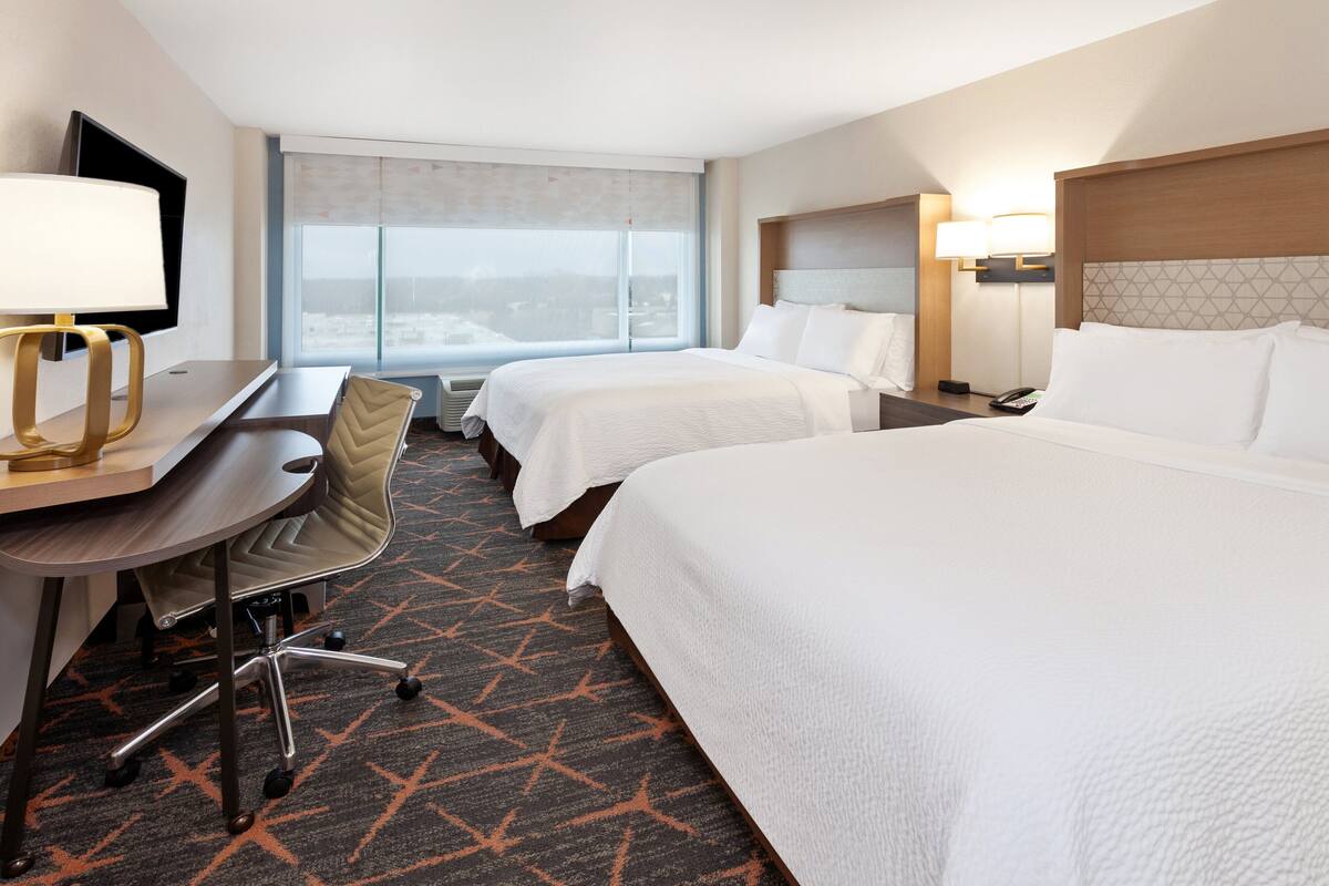 A hotel room with two beds, a desk with a chair, a TV, and a window with a view.