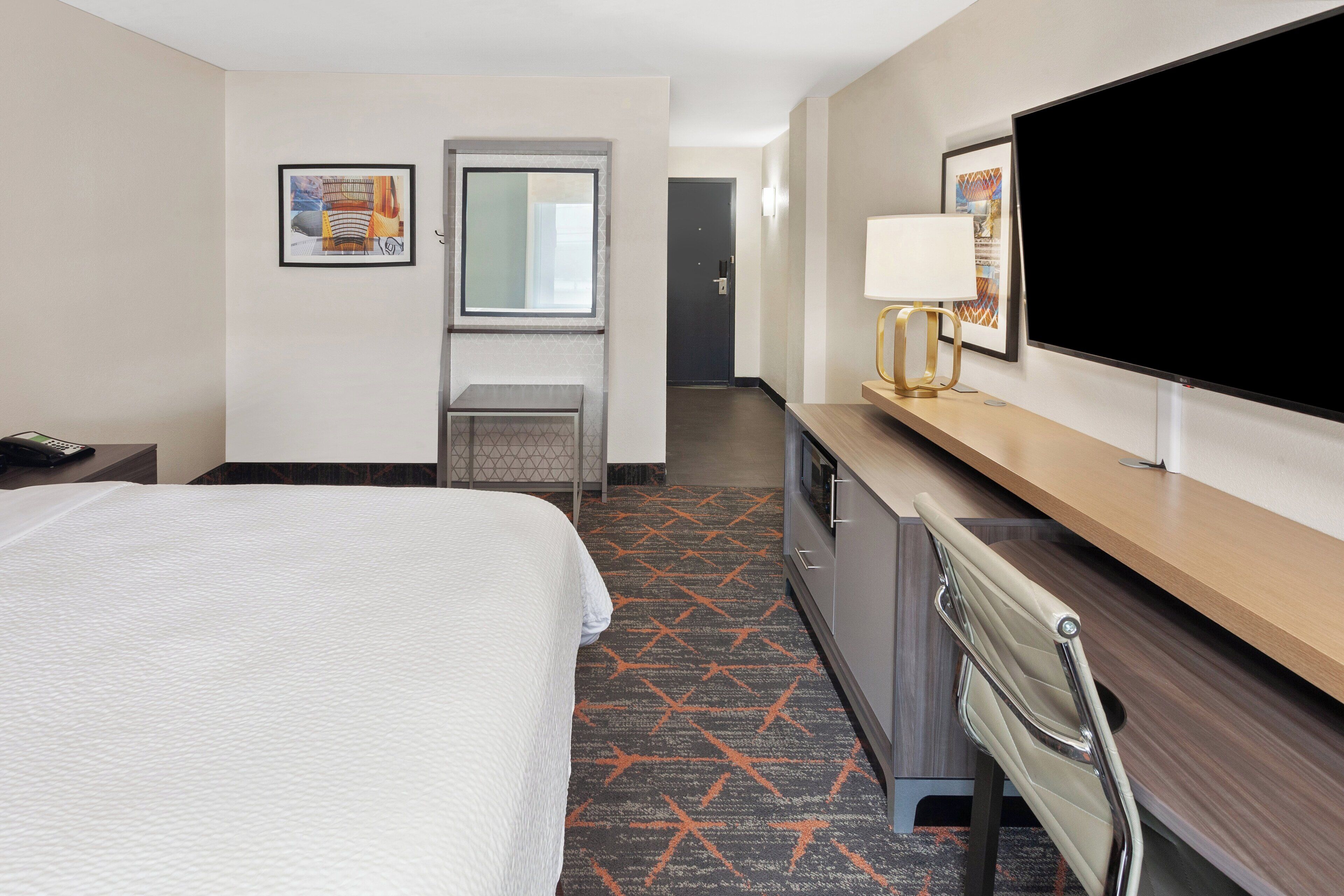A hotel room with a large bed, a desk with a flat-screen TV, a chair, a desk lamp, and a door leading to another room.