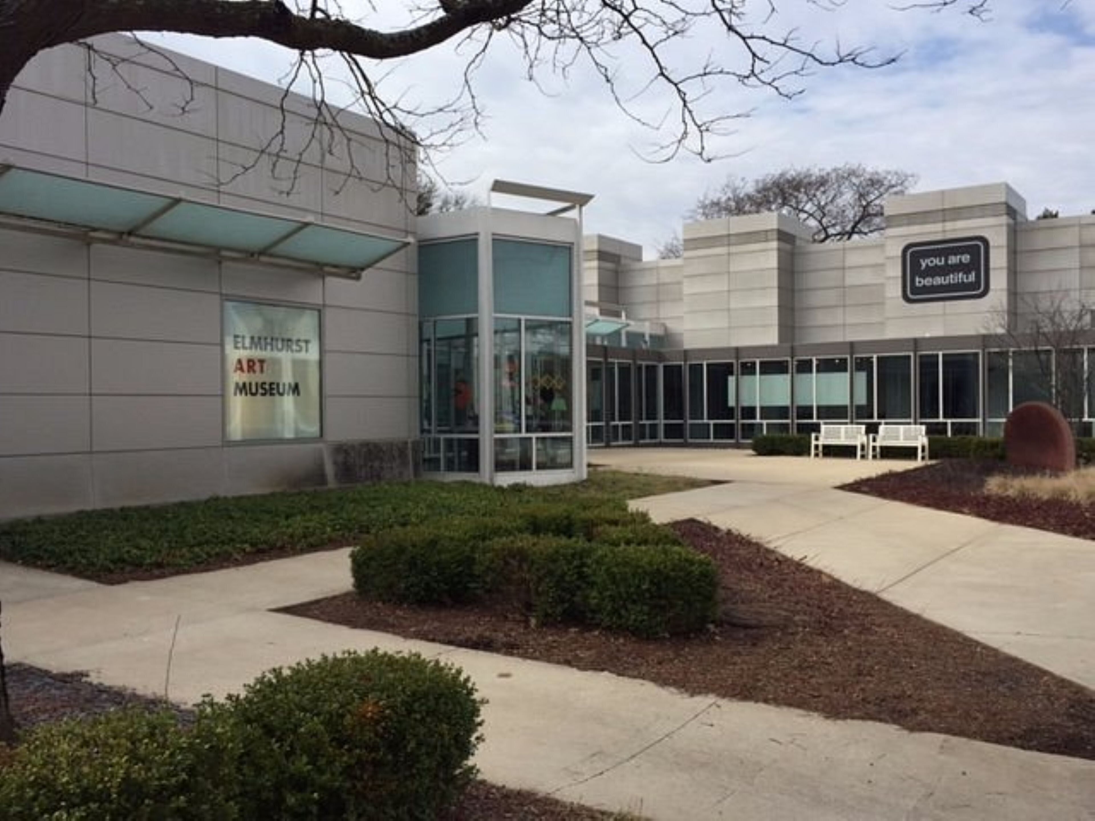 A modern building with a glass entrance, a sign reading 'Elmhurst Art Museum,' and a bench in the foreground.