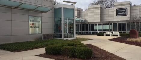 A modern building with a glass entrance, a sign reading 'Elmhurst Art Museum,' and a bench in the foreground.