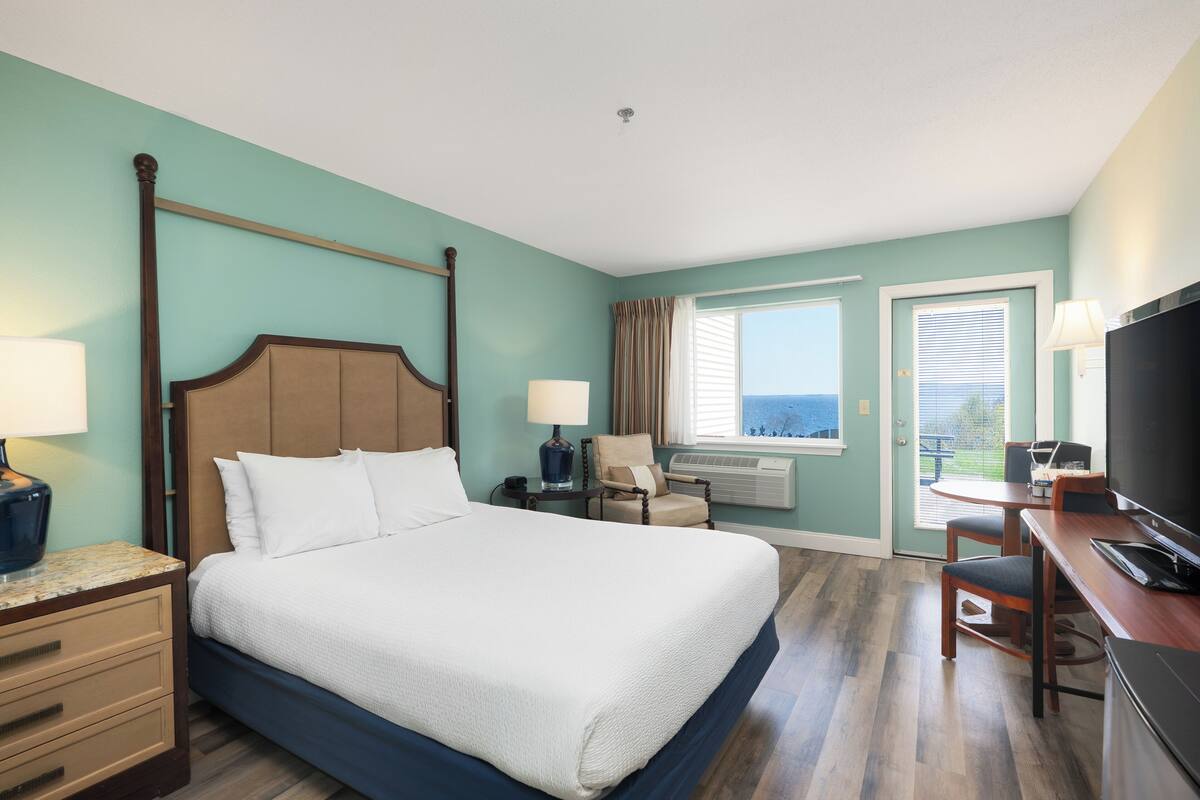 A hotel room with a four-poster bed, a desk with a computer, a chair, a TV, and a window with a view.
