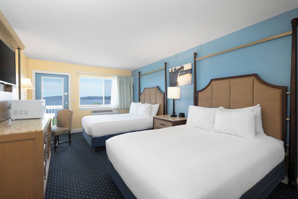 A hotel room with two beds, a desk with a microwave, a chair, a window with curtains, and a view of the outdoors.
