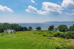 A grassy field with several white chairs arranged for an event, a house, and a view of the sea and distant hills.