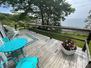 A wooden deck with turquoise chairs and a table, overlooking a body of water and a grassy area.
