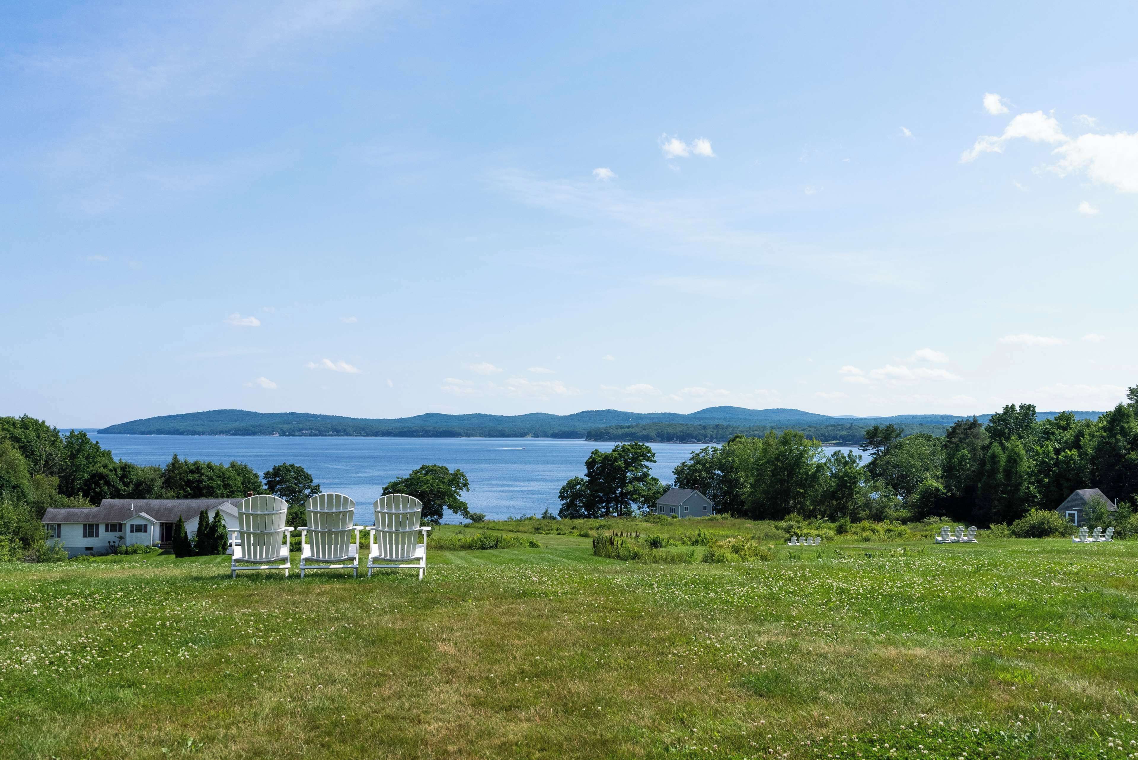 Un campo cubierto de hierba con sillas Adirondack blancas y vistas a un lago y montañas.