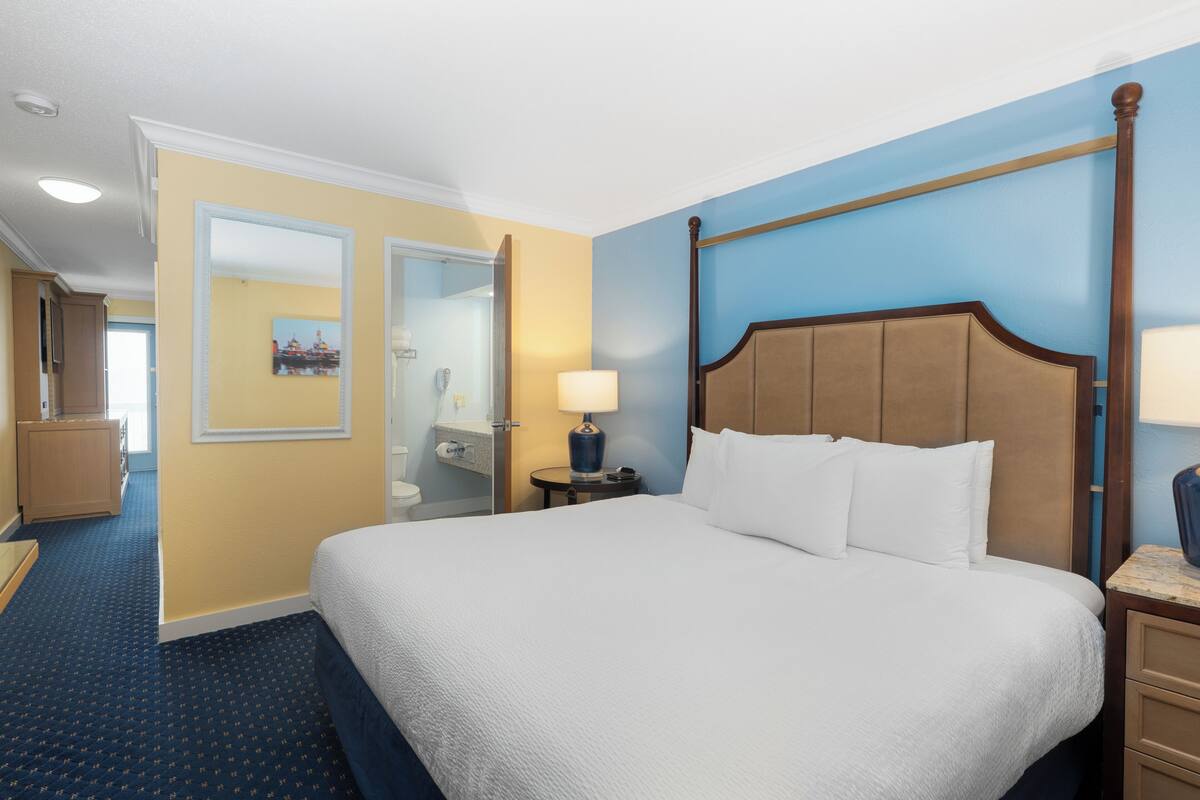 A hotel room with a large bed, a nightstand with a lamp, a wooden headboard, and a bathroom visible through an open door.