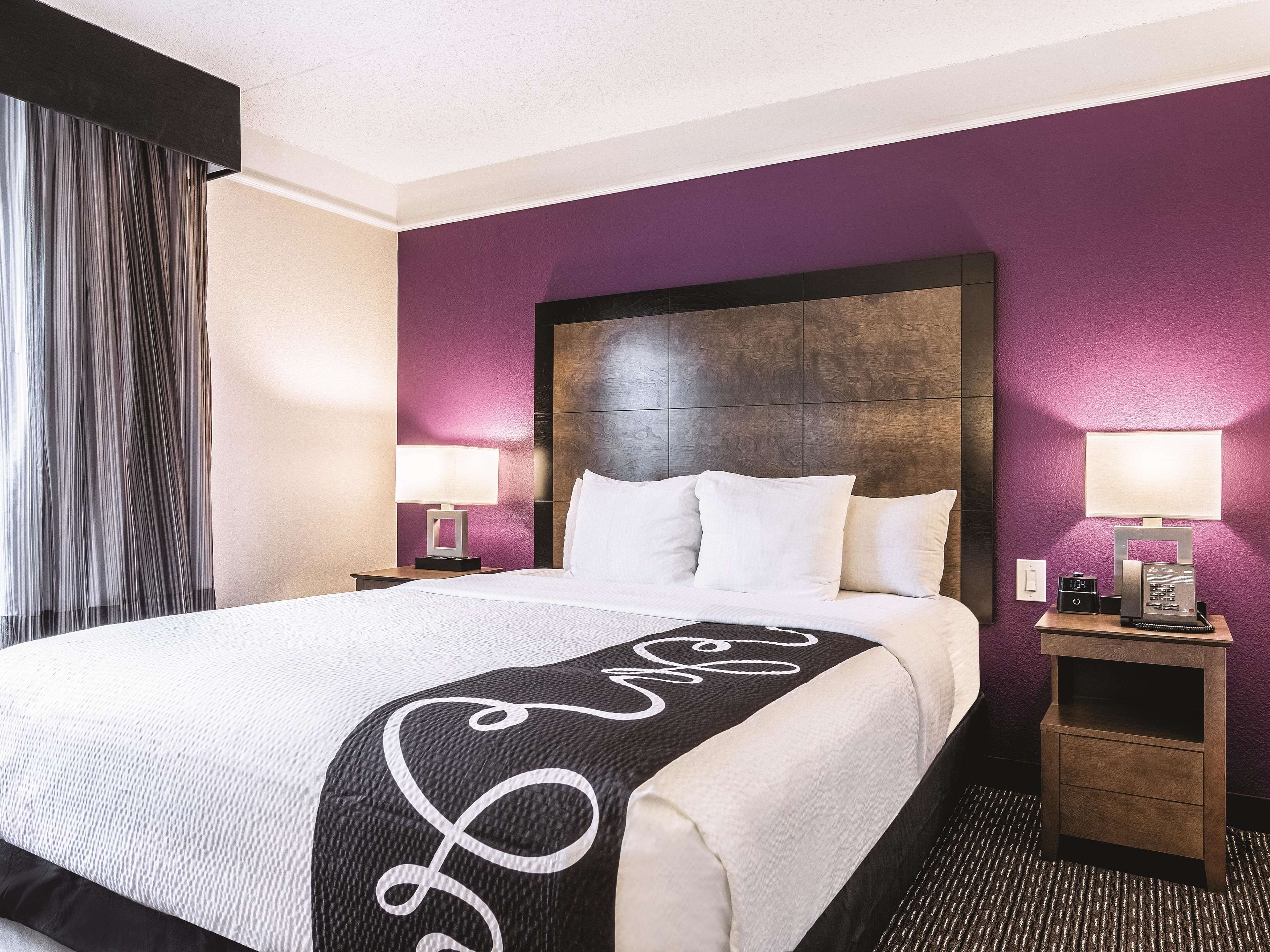 Ein Hotelzimmer mit einem großen Bett, violetten Wänden, einem hölzernen Kopfteil, zwei Nachttischen mit Lampen und einem Beistelltisch mit Telefon.