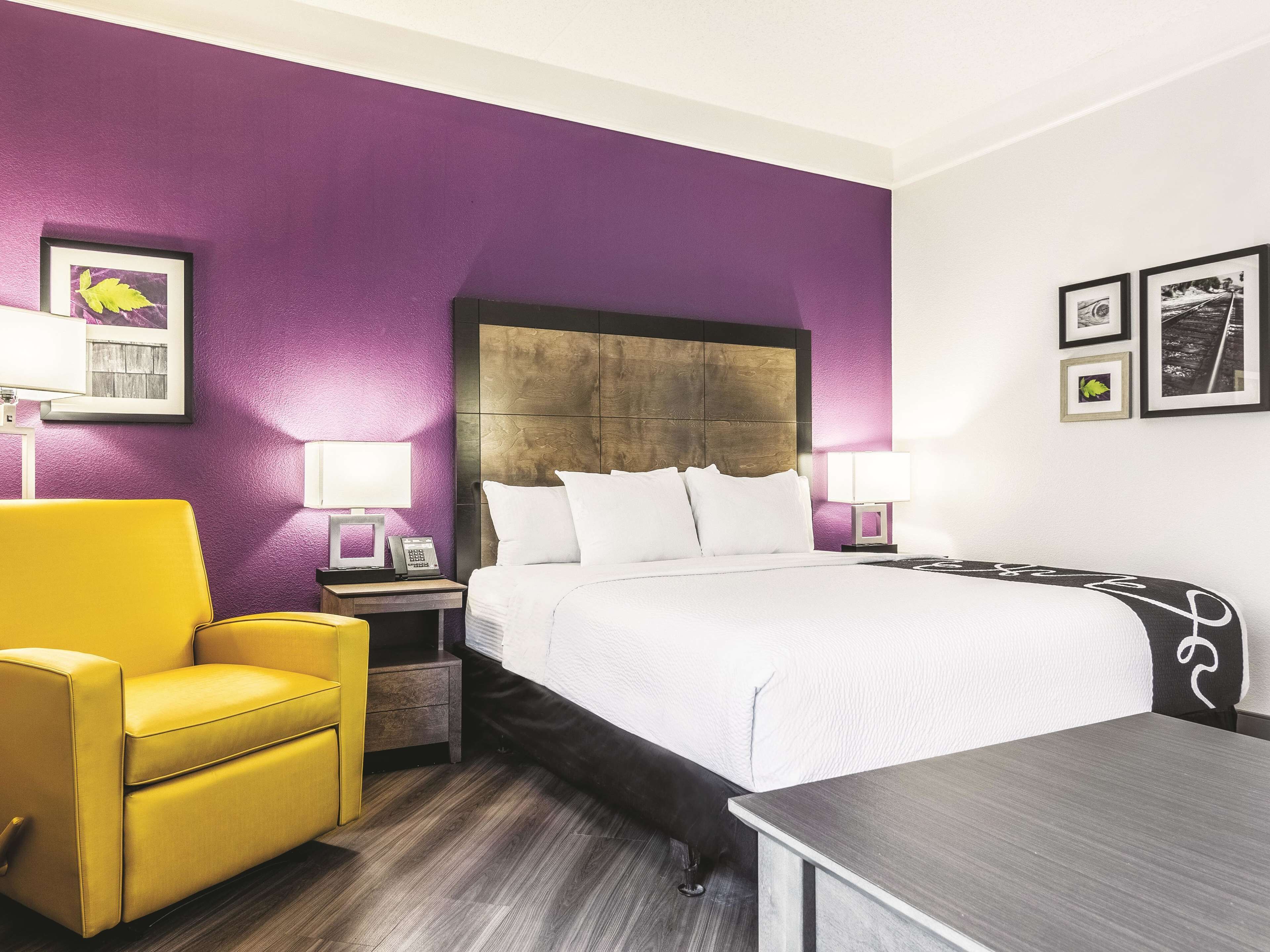 A hotel room with a large bed, a yellow armchair, a bedside table with a lamp, and a small table with a chair.