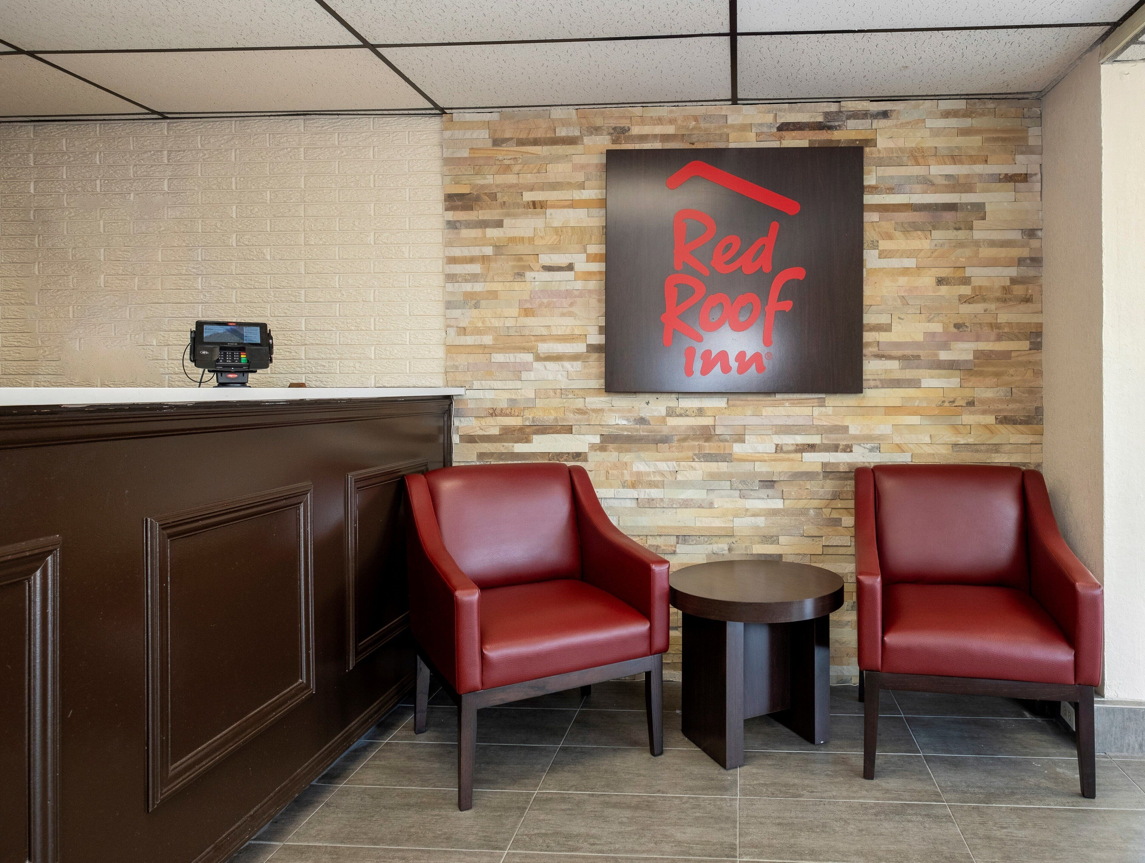 Área de recepção do Red Roof Inn, com parede de tijolos, balcão de recepção e duas poltronas de couro vermelho.