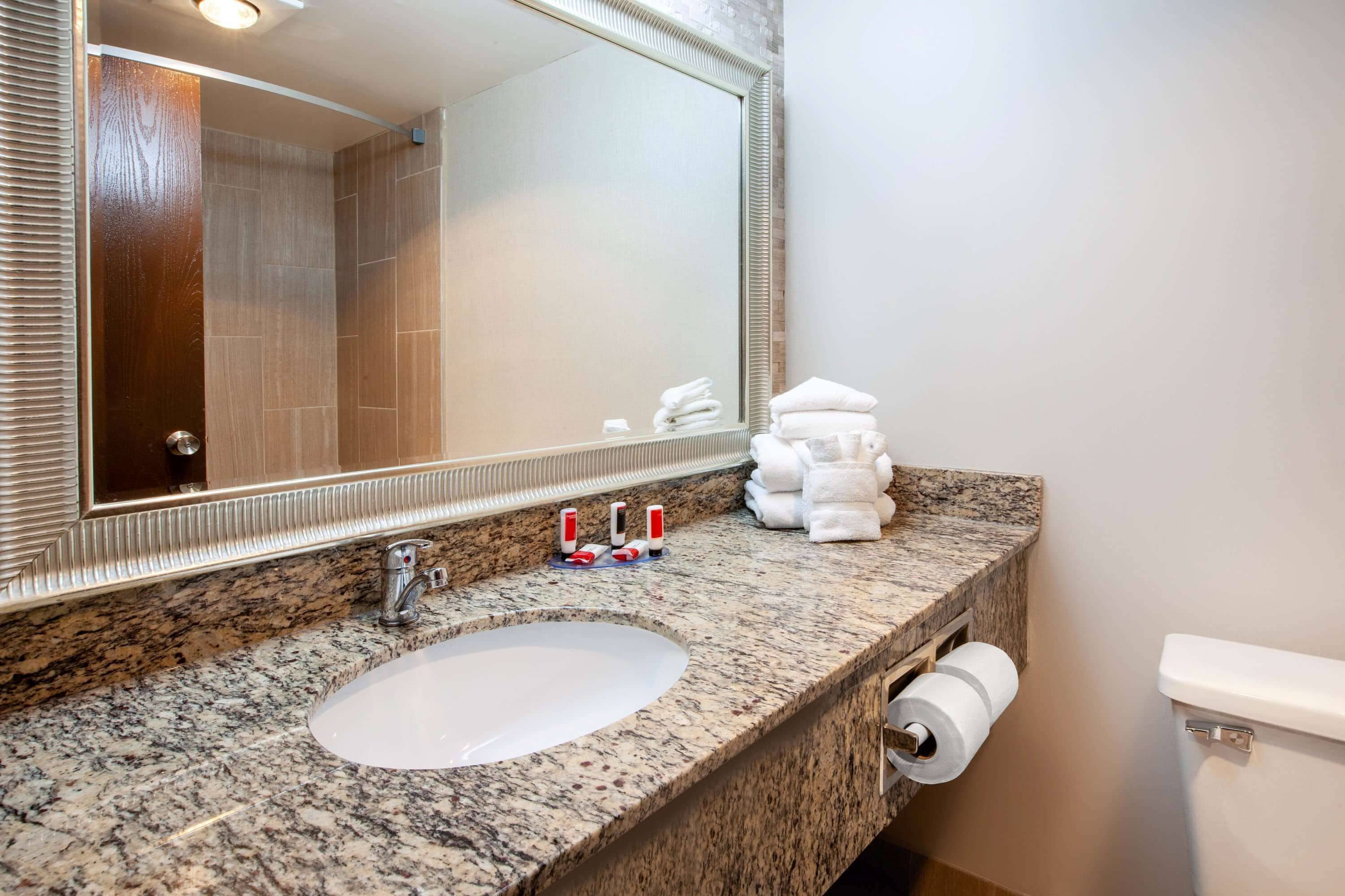 A bathroom with a granite countertop, a sink, a toilet, a large mirror, and a towel dispenser.